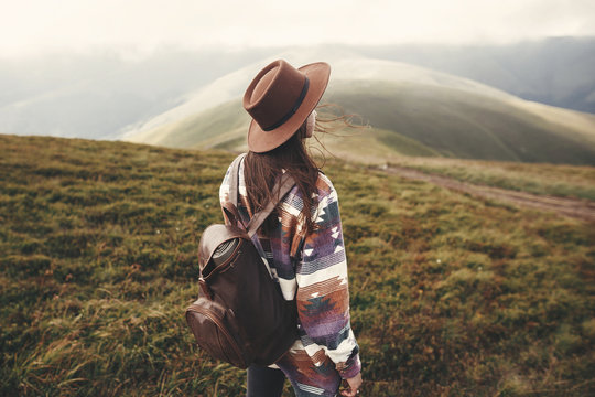 Stylish hipster girl in hat walking on top of mountains. Happy young woman with backpack exploring sunny mountains. Travel and wanderlust concept. Amazing atmospheric moment
