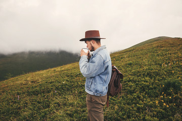 Stylish hipster traveler  drinking tea and standing on top of hills on  background of foggy mountains. Copy space. Handsome man in hat with backpack traveling in mountains. Wanderlust