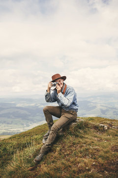 Stylish hipster traveler holding photo camera and taking picture on top of hills on background of foggy mountains. Copy space. Handsome man in hat with backpack traveling in mountains.