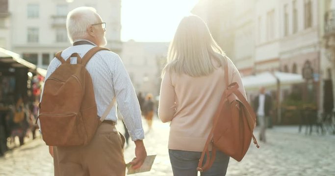 Back view on the senior Caucasian couple of tourists with backpacks walking the city street and talking on a sunny day. Outdoors. Rear.