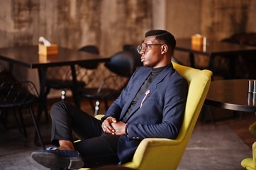 Fashionable african american man in suit and glasses sitting on chair at cafe.