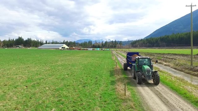 green farm tractor fertilizing large pece of farm land in beautiful BC surrounded by mountains and trees - shot from aerial drone in 60 frame per second