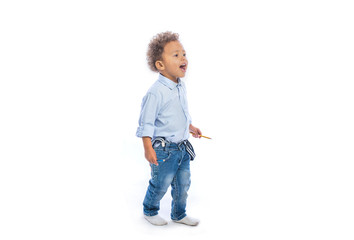 A little dark-skinned boy with curly hair in jeans and a light shirt stands in profile holding a pencil in his hands looking to the side and calling for someone