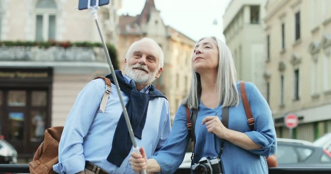 Caucasian senior smiled and happy man and woman tourists standing in the city outdoor with a smartphone on the selfie stick and videochatting ot taking photos.
