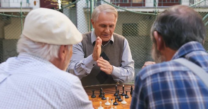 Caucasian old man happily doing a chessman move and being cheerful as having succeessful game. Rear of two other men. Outdoor.