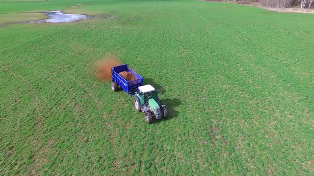 green farm tractor fertilizing large pece of farm land in beautiful BC surrounded by mountains and trees - shot from aerial drone in 60 frame per second