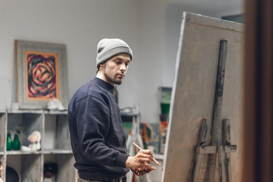 Busy man artist with a brush in his hand picks up an oil painting in a cozy studio. Talented young painter paints a painting on canvas. Painting Concept. Work as a hobby.