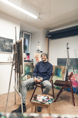 Happy painter sits on a chair near an easel with a canvas in his own studio, looks at the camera and smiles. Portrait of a positive young artist in the studio. Work as a hobby.