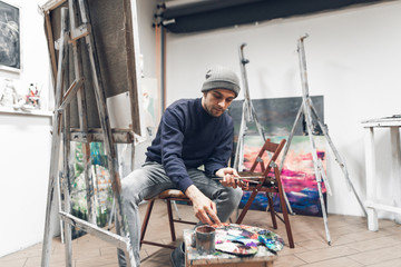 Naklejka na ściany i meble Concentrated professional painter with a brush in his hands is working on a painting in his own art studio, mixing paint and painting on canvas. Painting artist is in the work. Painting Concept.
