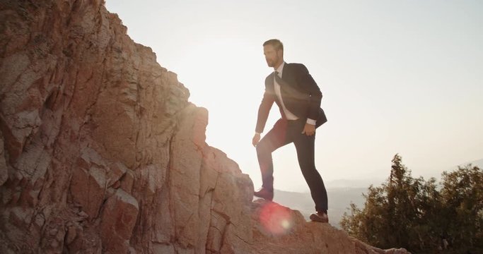 Young confident businessman climbing up the rocky mountain, wearing suit and tie - way to success concept 4k