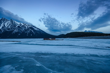 Lake Minnewanka Banff, Canada. Glacial lake completely frozen and covered in snow. Blue hour at the lake with glacial mountain background. Cabin on the frozen lake.