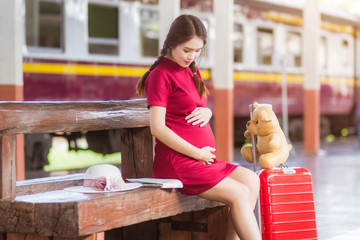 Asian woman pregnant red dress sitting on a bench at carrying red luggage and Teddy Bear at railway station travel.