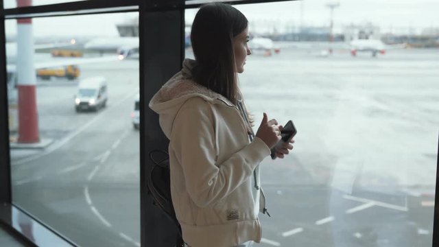 Woman uses mobile phone and waves her hand to someone from window at airport.