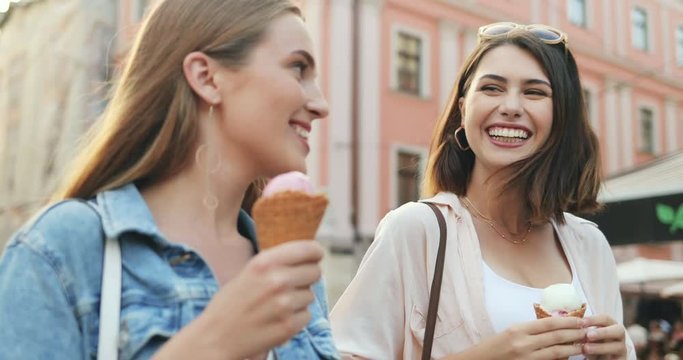 Close up of the two joyful young Caucasian women friends walking the city center, talking and eating ice-cream. Outdoors.
