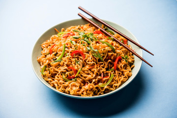Schezwan veg noodles is a spicy and tasty stir fried flat Hakka noodles with sauce and veggies. served with chopsticks. selective focus