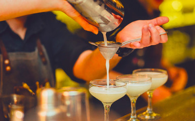 Cocktail being poured at a bar preparing a drink. Motion blur of the bartender's hand and strainer. Selective focus on the foreground glass. Blurred people in the background.  Trendy black stylish 