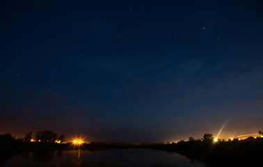  Lake under the stars. Pond on the outskirts of the city. Night summer sky. City lights in the night. Blue starry sky. The lights are far away.