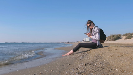 Fototapeta na wymiar Traveler girl with a backpack sitting on a sandy sea beach and typing a message on mobile phone.