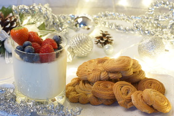 Panakota with berries in a glass, near a stack of cookies, garlands and spangles on a light background, Christmas mood