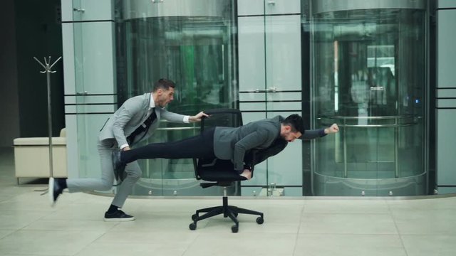Playful business people are having fun in office lobby riding chair and playing racing game on rolling armchair. Emotional youth, joyful adults and work concept.