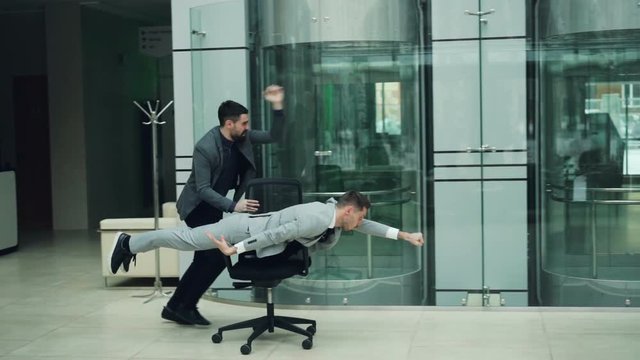 Bearded guys businessmen are having great time at office party riding chair in lobby enjoying freedom amd good mood. Funny adults and workspace concept.