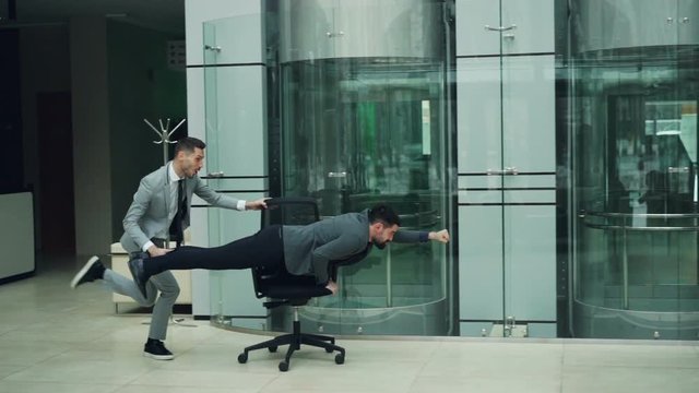Joyful office workers are having fun with rolling chair in lobby of business center riding armchair and laughing enjoying funny activity at work party.