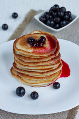 Stack of american pancakes with blackcurrant jam and fresh blackcurrant berries are lying on a white plate.