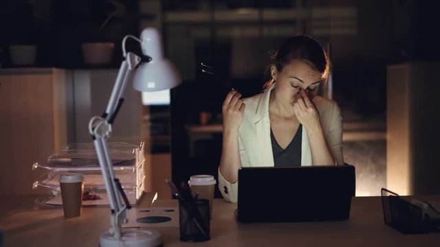 Beautiful girl in formal clothing in working on laptop alone in dark office late at night finishing urgent work overworking. Modern technology and job concept.