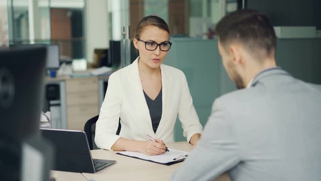 Friendly businesswoman in glasses and suit is interviewing a male candidate for job in office. People are talking asking and answering questions, girl is writing.