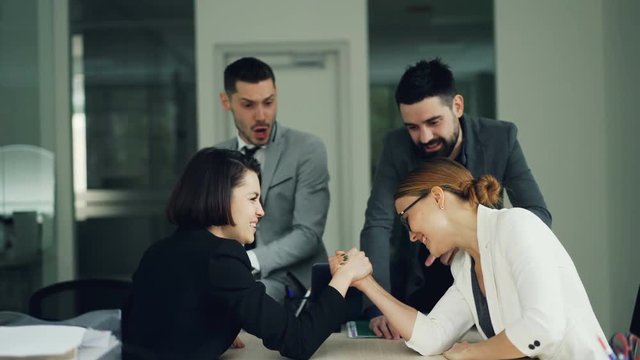 Cute girls office workers are competing in arm wrestling sitting in room at desk while young men in suits are cheering them then doing high-five. Fun and job concept.