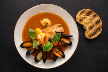 Seafood fish soup with mussel and shrimps and piece of grilled bread on black background
