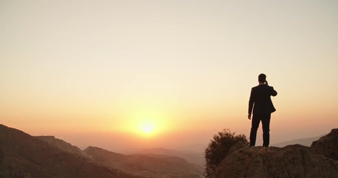 Young confident businessman in white collar standing on top of the mountain, looking far away and talking on phone - way to success concept 4k