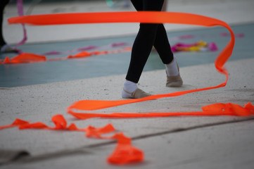 Spirals with orange art ribbon in fitness class. Young little gymnast girl in black sportswear dress, doing rhythmic gymnastics exercise with orange ribbon