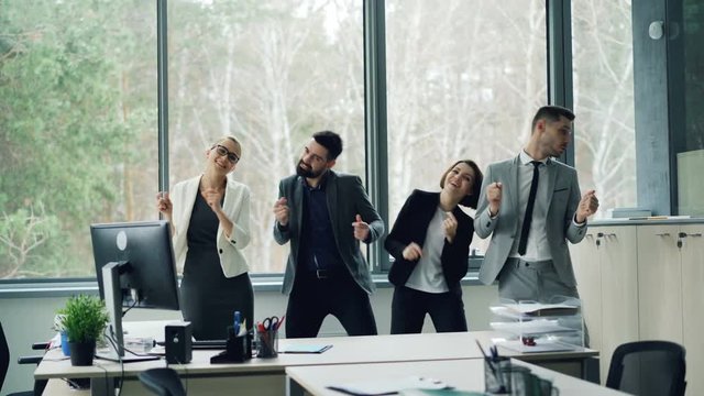 Joyful team of young coworkers is dancing in modern office wearing suits moving arms and body smiling and laughing. Relaxing at work and partying concept.