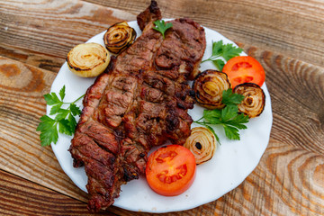 Grilled pork meat with onion, tomato and parsley in white plate on wooden table
