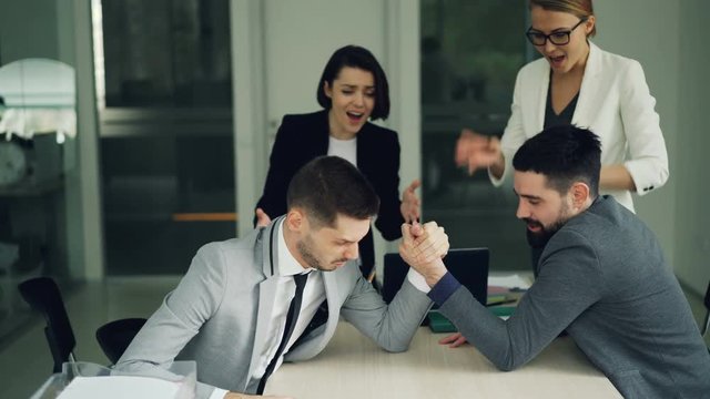 Bearded young people office workers are having fun practising arm wrestling while women are watching and supporting them then doing high-five and clapping hands.