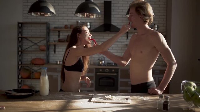 Young sexy hot girl in bra and handsome man in the kitchen feed each other hot chilli red pepper. Loving couple in underwear having fun in the kitchen. Slow motion.