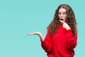 Beautiful brunette curly hair young girl wearing glasses and winter sweater over isolated background amazed and smiling to the camera while presenting with hand and pointing with finger.