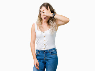 Fototapeta premium Young beautiful blonde woman over isolated background peeking in shock covering face and eyes with hand, looking through fingers with embarrassed expression.