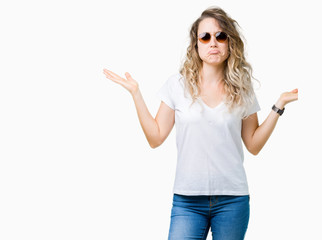 Beautiful young blonde woman wearing sunglasses over isolated background clueless and confused...