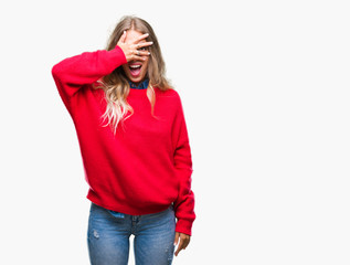 Beautiful young blonde woman wearing winter sweater over isolated background peeking in shock covering face and eyes with hand, looking through fingers with embarrassed expression.