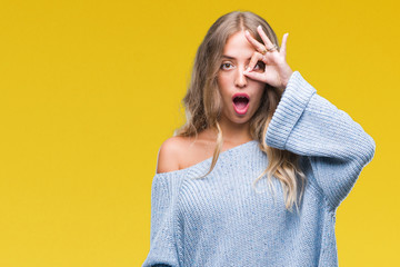 Beautiful young blonde woman wearing winter sweater over isolated background doing ok gesture shocked with surprised face, eye looking through fingers. Unbelieving expression.