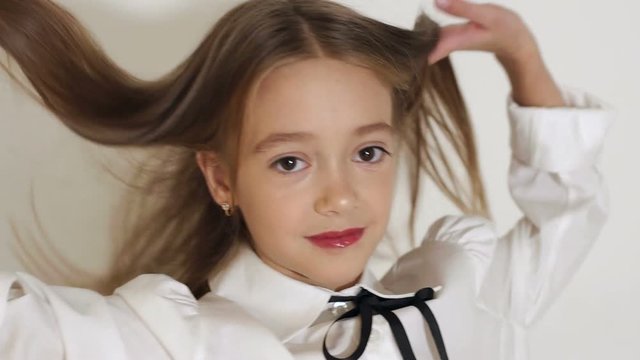 Close-up portrait of a cute little girl with big brown eyes and long hair posing in front of the camera in a studio with a white wall, she looks into the camera and throws up her hair with her hands.