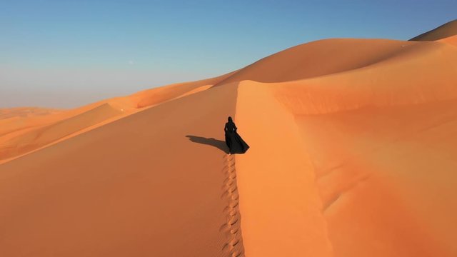  Aerial view from a drone flying next to a woman in abaya (United Arab Emirates traditional dress) walking on the dunes in the desert. Dubai, UAE.