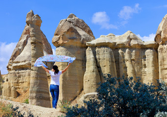 A young woman stands in the wind between huge, old, conical and weathered rocks in the honey valley of Cappadocia under the bright day sun.