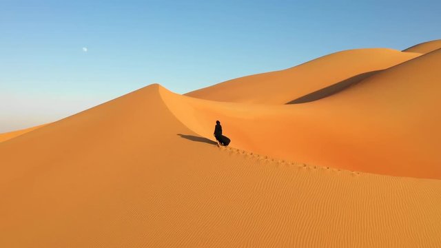  Aerial view from a drone flying next to a woman in abaya (United Arab Emirates traditional dress) walking on the dunes in the desert. Dubai, UAE.