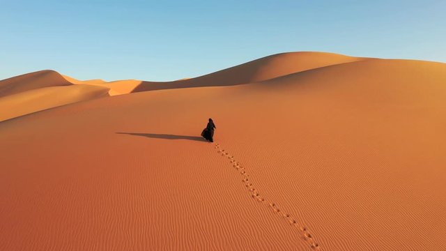  Aerial view from a drone flying next to a woman in abaya (United Arab Emirates traditional dress) walking on the dunes in the desert. Dubai, UAE.