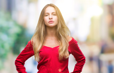 Young beautiful blonde woman wearing red sweater over isolated background with serious expression on face. Simple and natural looking at the camera.