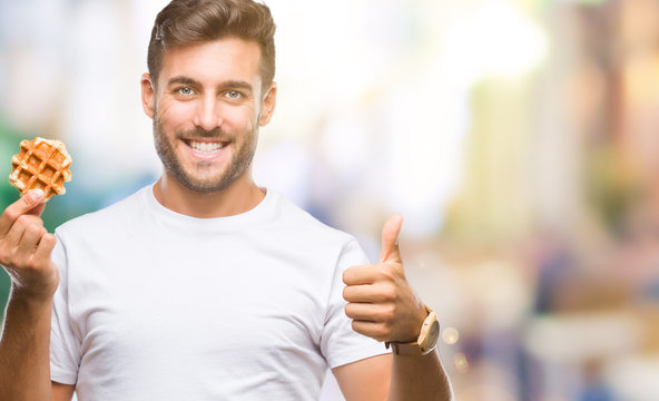 Young handsome man eating a sweet waffle over isolated background happy with big smile doing ok sign, thumb up with fingers, excellent sign