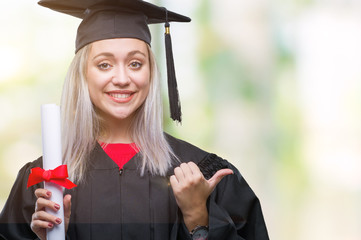 Young blonde woman wearing graduate uniform holding degree over isolated background pointing and...